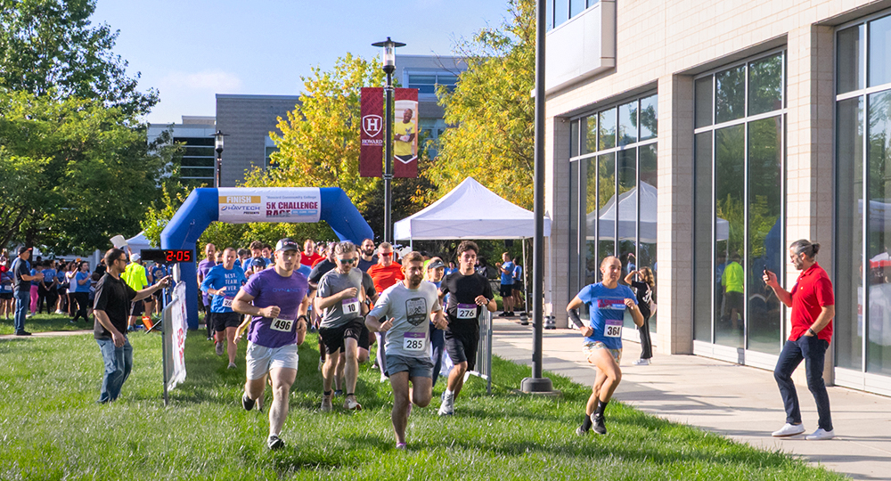 Runners take off from the starting line of the Howard Community College 5K Challenge Race, passing under a blue inflatable arch with a crowd of participants and spectators behind them.