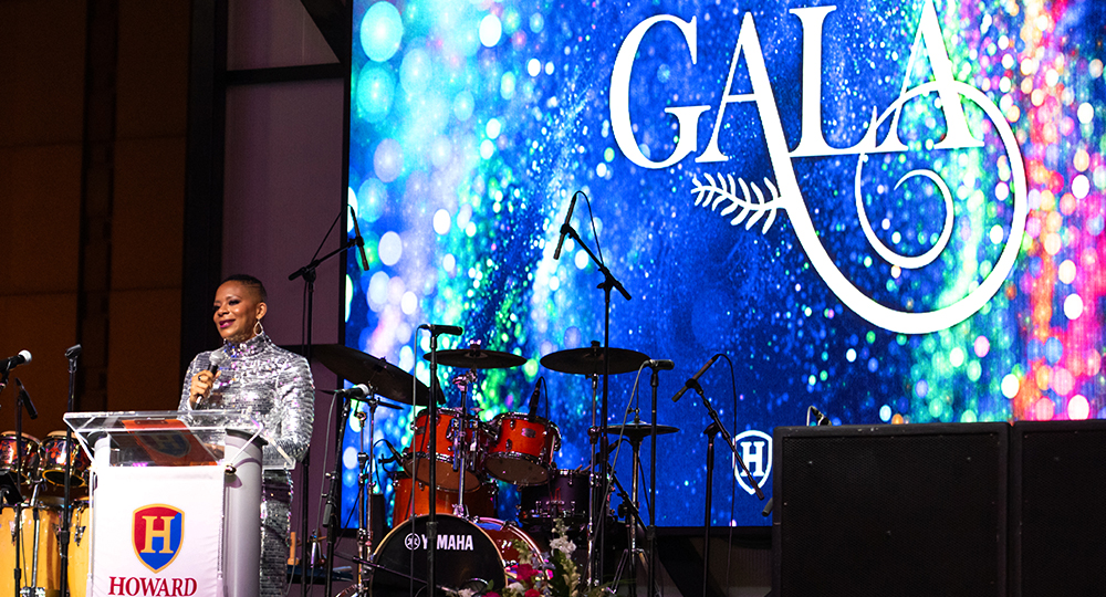Dr. Daria J. Willis stands at a podium with the Howard Community College logo, addressing the audience at a formal event. Behind them is a brightly lit stage with musical instruments and a large colorful screen that reads 