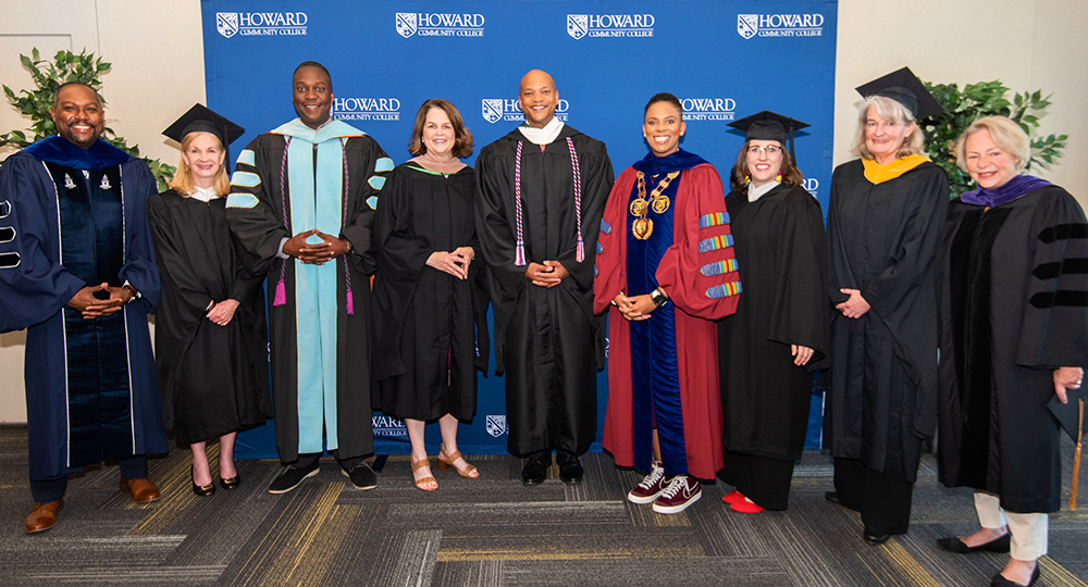 group of HCC staff and guests dressed in cap and gown standing in front of the blue step and repeat with HCC logos on the fabric