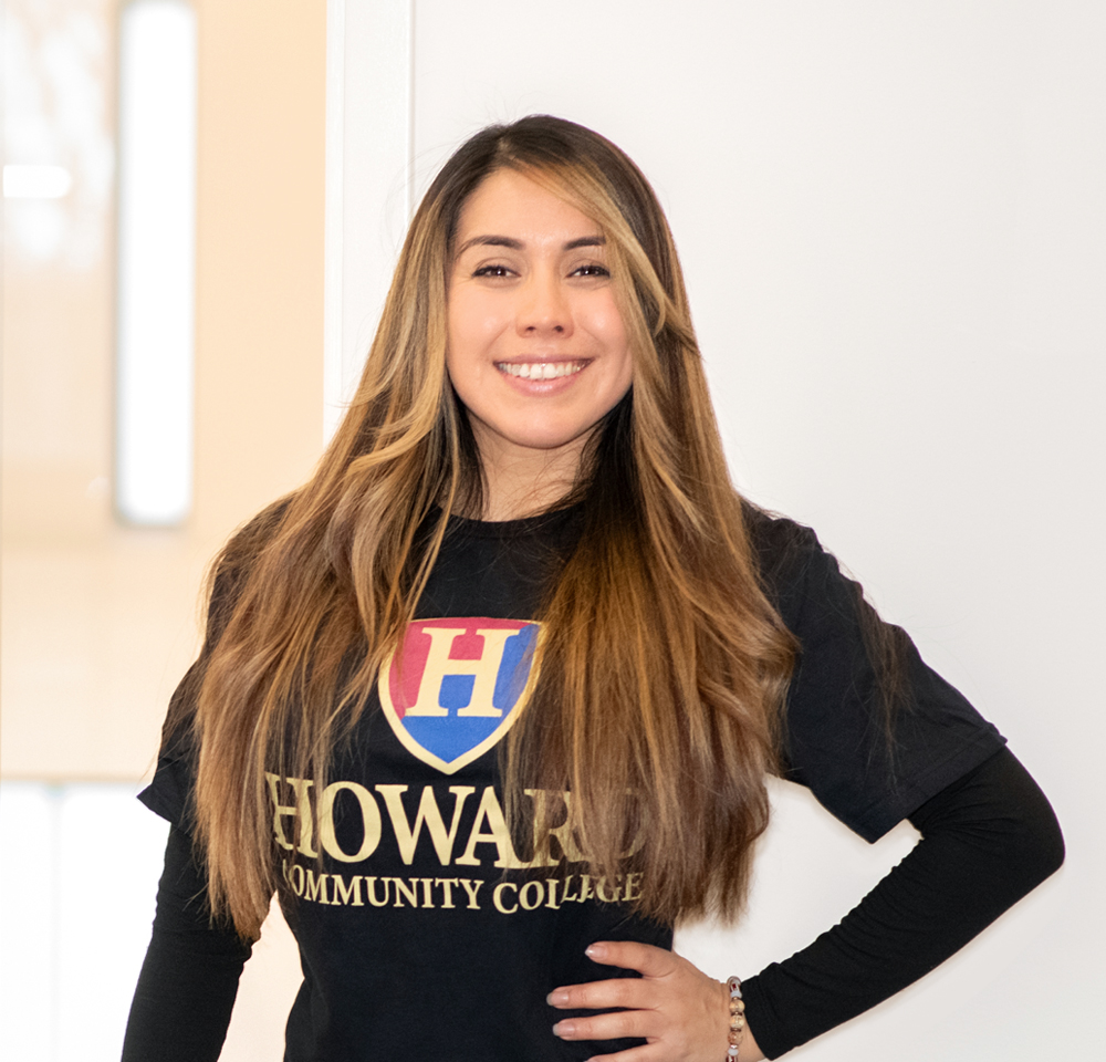 Dayana Paez Ortiz is a a young woman with long, wavy, light brown hair and a smile stands against a white wall. She is wearing a black Howard Community College T-shirt with the school's logo in red, blue, and gold on the front. Her left hand is on her hip, and her right arm hangs naturally by her side. She has a warm and confident expression.
