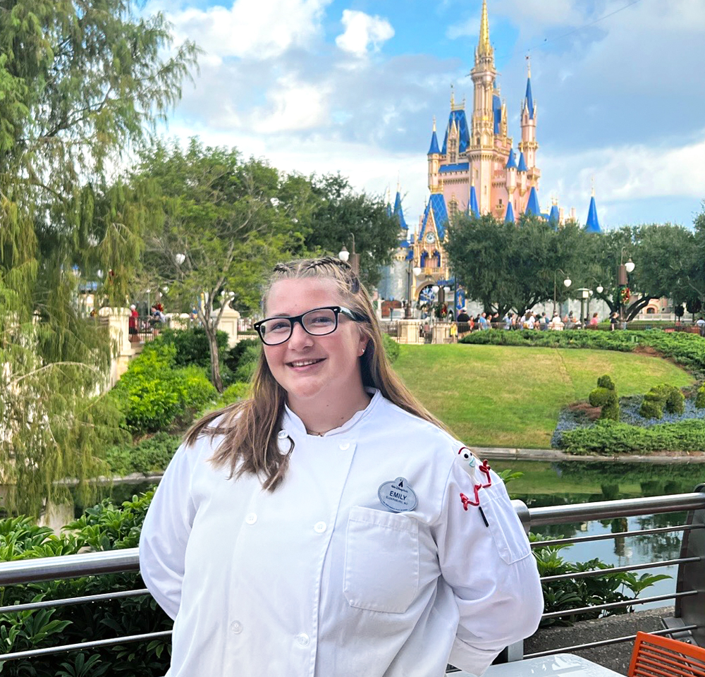 A young woman wearing a white chef's coat and black glasses smiles while standing in front of Cinderella Castle at Walt Disney World. Her name tag reads “Emily,” and a small Forky character from Toy Story is embroidered on her sleeve. Lush greenery, a waterway, and the iconic castle are visible in the background under a partly cloudy sky.