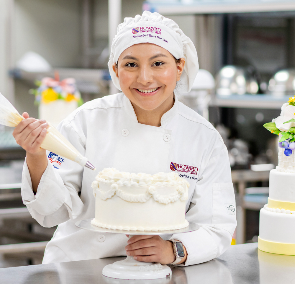 A smiling young woman in a white chef's uniform and hat is decorating a white frosted cake with a piping bag in a professional kitchen. Her uniform features a logo for Howard Community College. She holds the cake on a stand and appears to be mid-decoration.