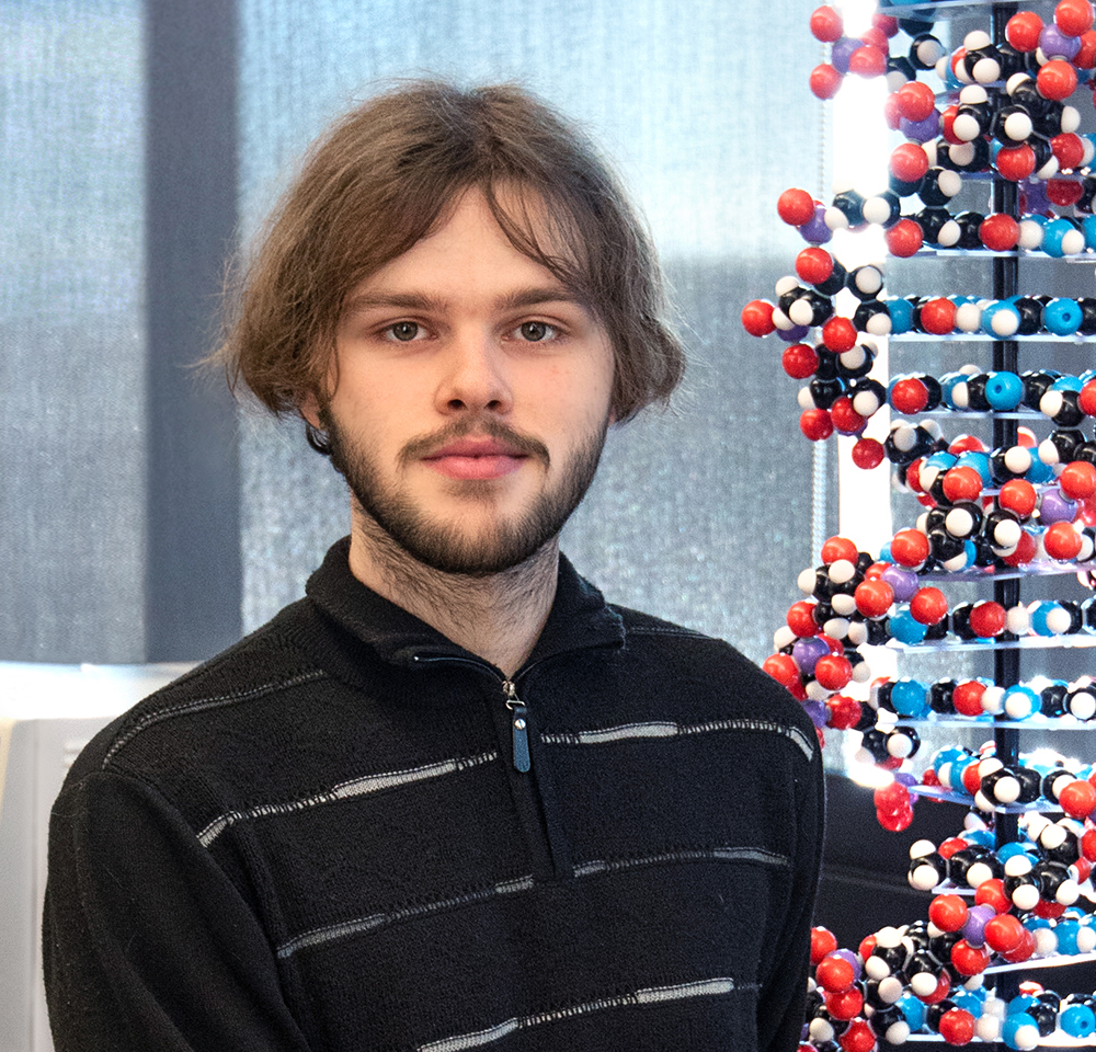 A young man with medium-length brown hair and a beard is standing indoors in front of a large 3D model of a DNA double helix. He is wearing a black zip-up sweater with light gray stripes and is looking directly at the camera with a slight smile. The background includes dark curtains and bright lighting on the DNA model.