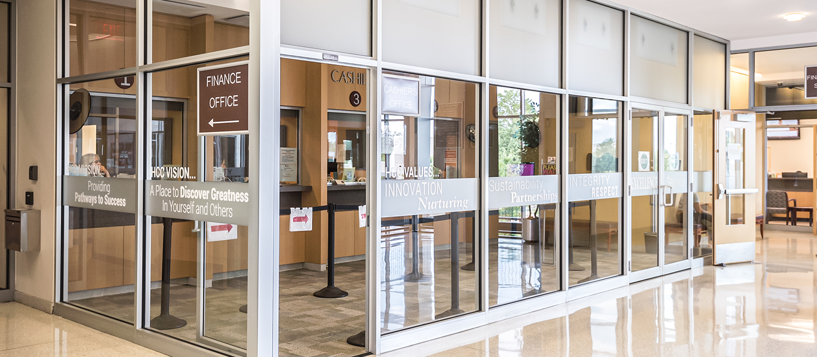 A glass-walled college finance and cashier’s office with multiple service windows, interior wood paneling, and directional signage. Words such as “Finance Office,” “Cashier’s Office,” and values like “Innovation,” “Integrity,” and “Excellence” appear on the glass panels. The hallway outside has polished floors and bright natural light.