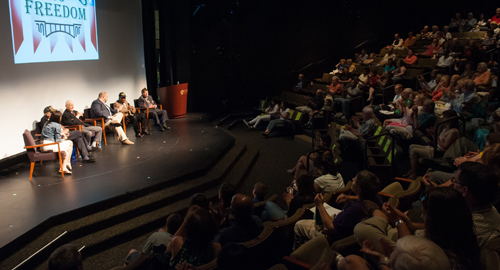 Panel discussion on a lit stage in a packed auditorium with a large projected banner