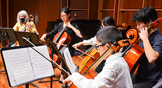 Five seated musicians on a wooden stage playing cellos and reading sheet music from stands; one musician at the left is wearing a face mask