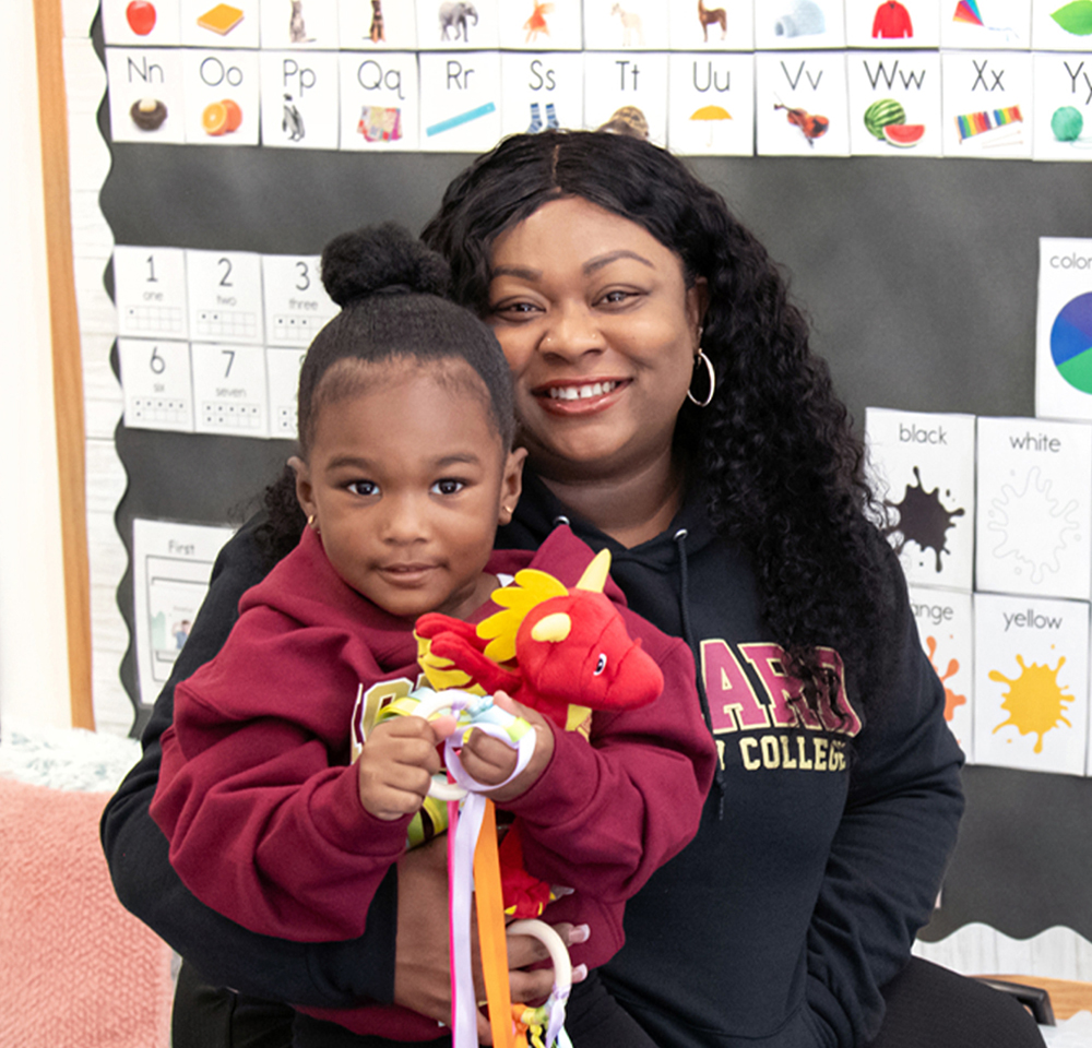 A smiling adult woman with long, curly hair and hoop earrings is holding a young child in her arms. Both are wearing maroon-colored hoodies, and the child is holding a red plush dragon toy with yellow accents and colorful ribbons. They are in a classroom with an alphabet chart and color labels on the wall behind them.