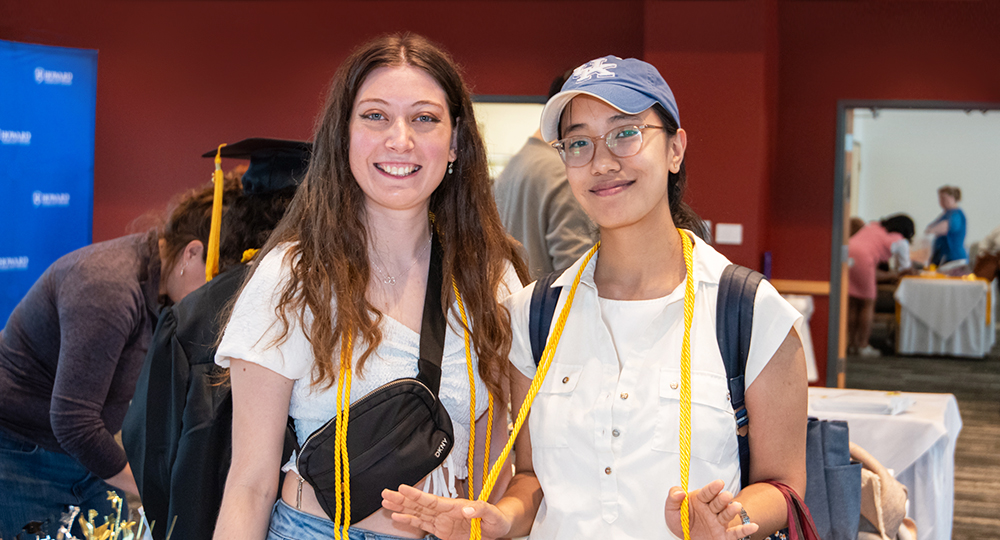 Two young women smile at the camera during a graduation event. Both are wearing honor cords around their necks. Other people and tables with graduation materials are visible in the background.