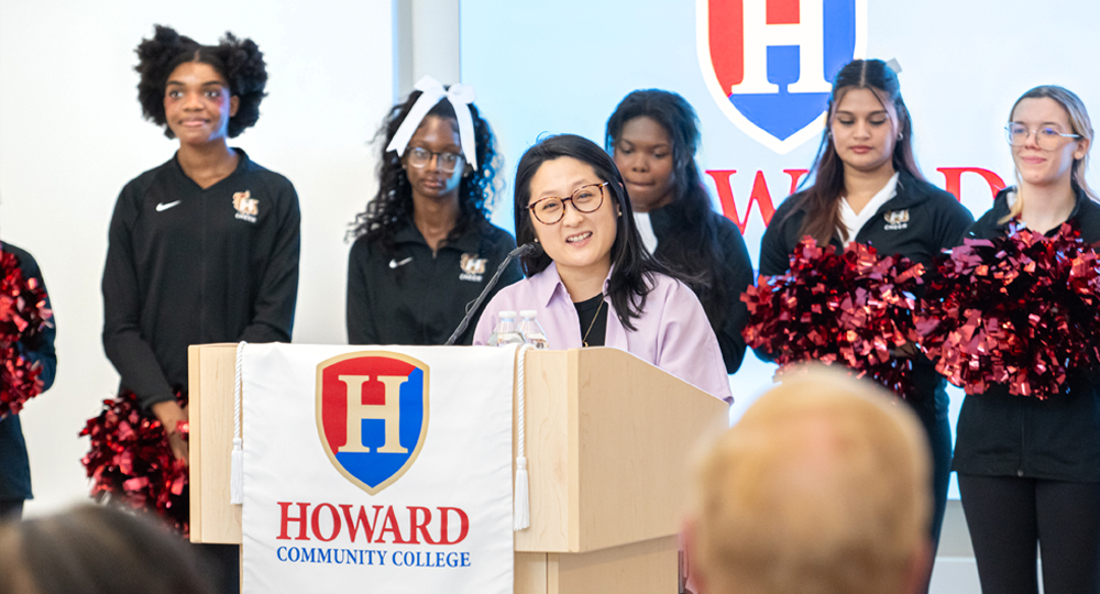 A woman speaks at a podium decorated with a Howard Community College banner, flanked by a group of cheerleaders holding red pom-poms. The cheerleaders are dressed in black uniforms, and a large Howard Community College logo is visible in the background.