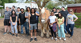 A diverse group of young people posing outdoors with a large light brown horse. They are standing on a wood-chip-covered area with trees, a white building, and a red barn in the background. Everyone is smiling and dressed casually.