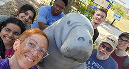 A group of seven people posing for a selfie with a large gray manatee statue. They are outdoors, smiling, and dressed casually. A sign and trees are visible in the background.