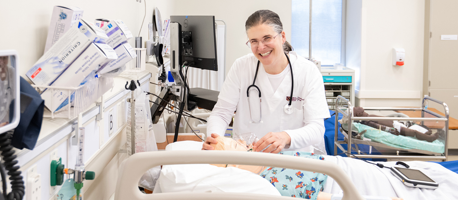 Smiling healthcare student in a white uniform and stethoscope practices patient care on a medical manikin in a clinical simulation lab, surrounded by medical equipment and monitors.