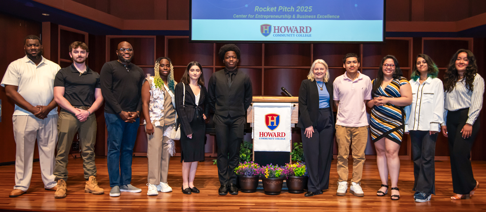 A diverse group of twelve individuals, including students and faculty, stands on a stage at Howard Community College in front of a podium and presentation screen that reads
