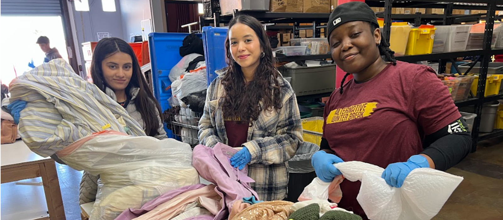 Students volunteering during an Alternative Winter Break, sorting donated clothing in a community service center. They smile while working together in a warehouse filled with bins and supplies.
