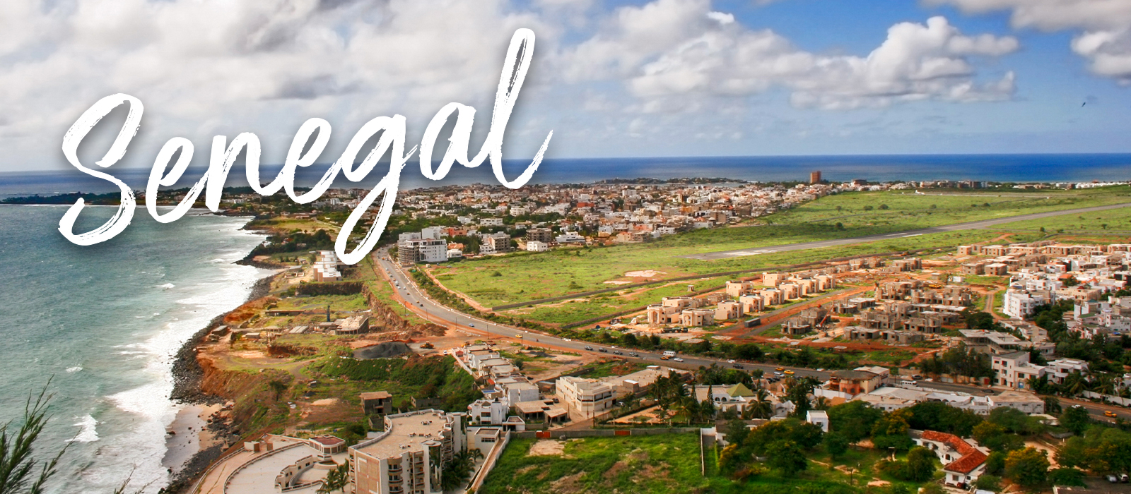 Aerial view of Dakar, Senegal, showing a coastal cityscape with buildings, green open land, and the Atlantic Ocean under a partly cloudy sky. The word “Senegal” is written in large white script across the image.