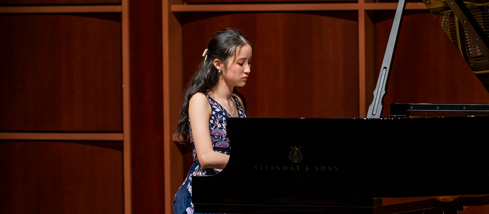 oung pianist seated at a Steinway & Sons grand piano, performing on a concert hall stage.
