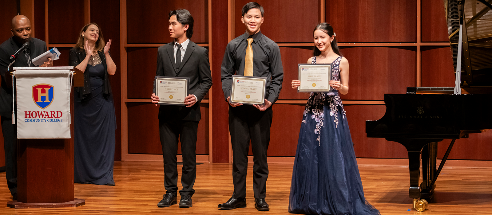 Student musicians receive competition awards on stage at a concert hall, standing beside a Steinway grand piano.
