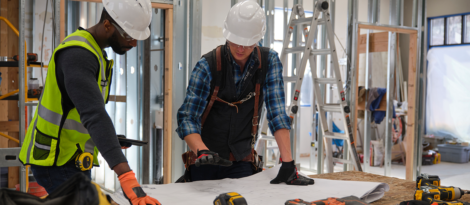 Two construction workers wearing safety gear and hard hats review blueprints inside a building under construction.