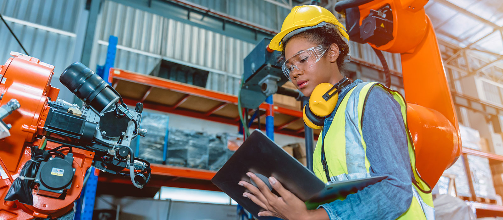 A worker in a hard hat and safety vest uses a laptop near an orange robotic arm in a warehouse, reflecting technology in industrial settings.
