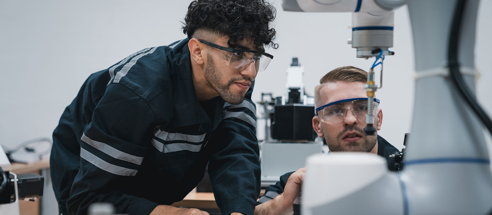 Two engineers in safety goggles focus intently on a robotic arm in a lab. One leans in with curiosity, conveying a sense of teamwork and innovation.