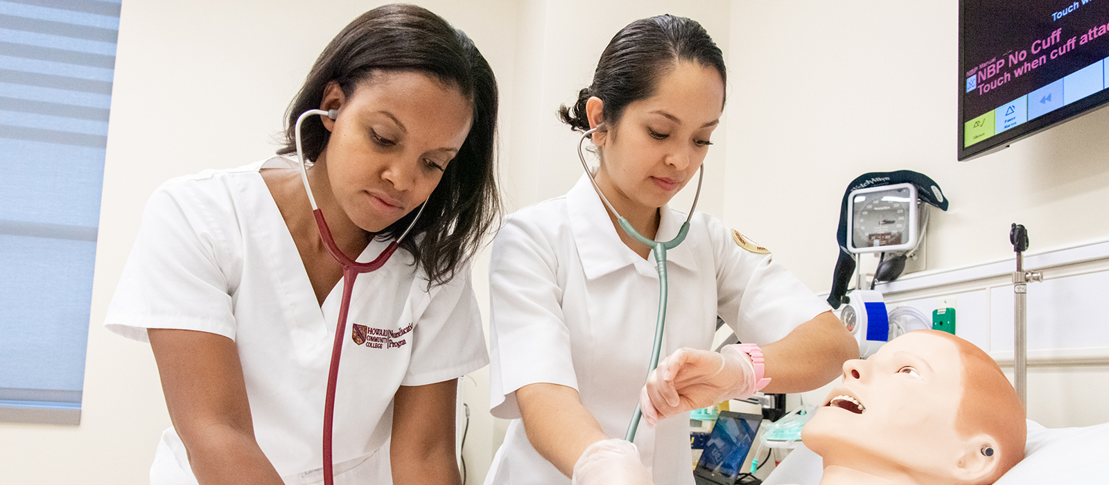 Two nursing students using stethoscopes to check the heart of a dummy lying in a hospital bed