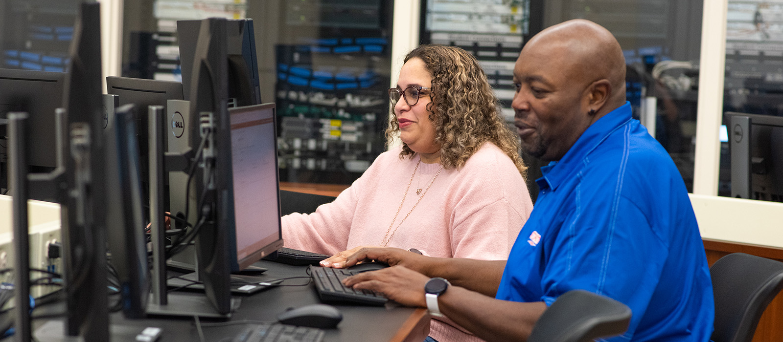 Two people sitting in front of a computer with servers and networking equipment in the background