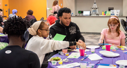 Students sit around a table covered with art supplies, engaging in a creative activity during a campus event.