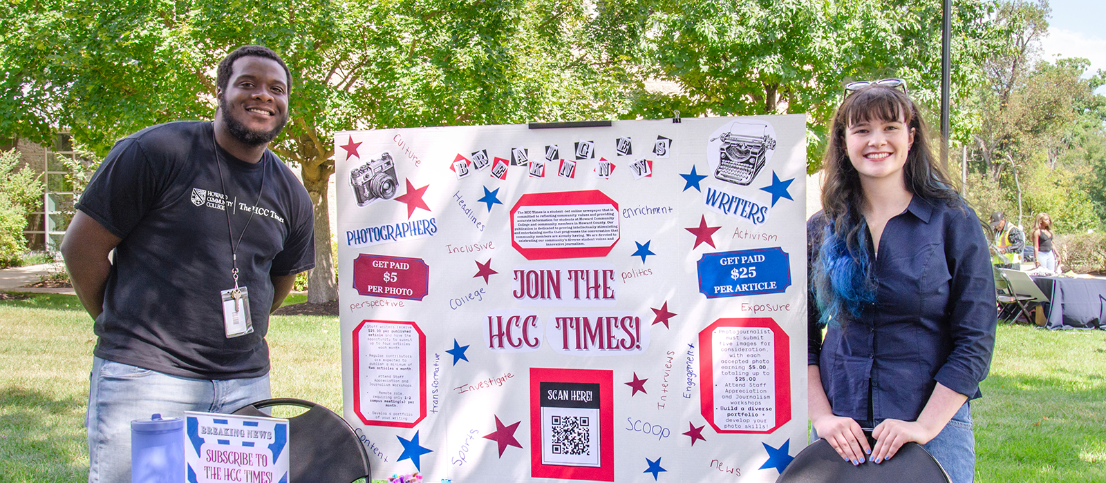 Two students stand outdoors beside a colorful poster board promoting The HCC Times student newspaper at Howard Community College.